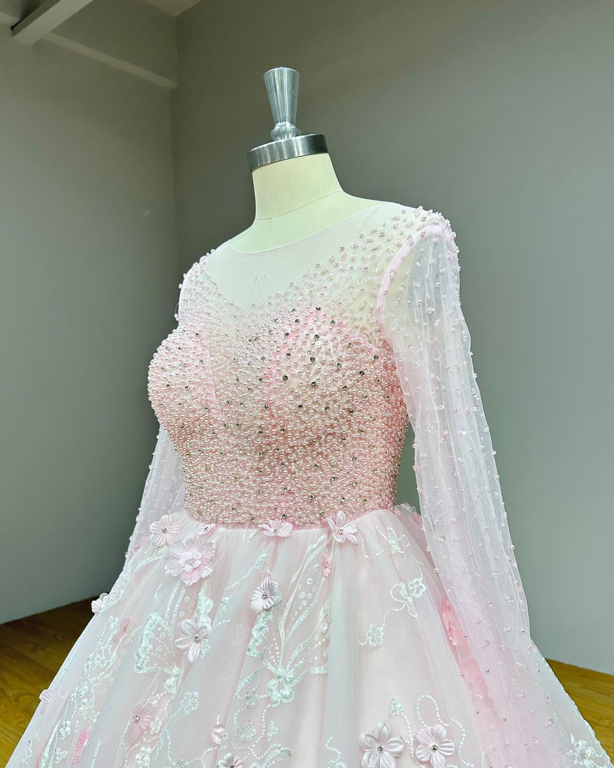 Side profile detail of a structured pink bridal bodice showing the intricate crystal beadwork pattern, sheer long sleeves, and 3D floral appliques at the waistline.