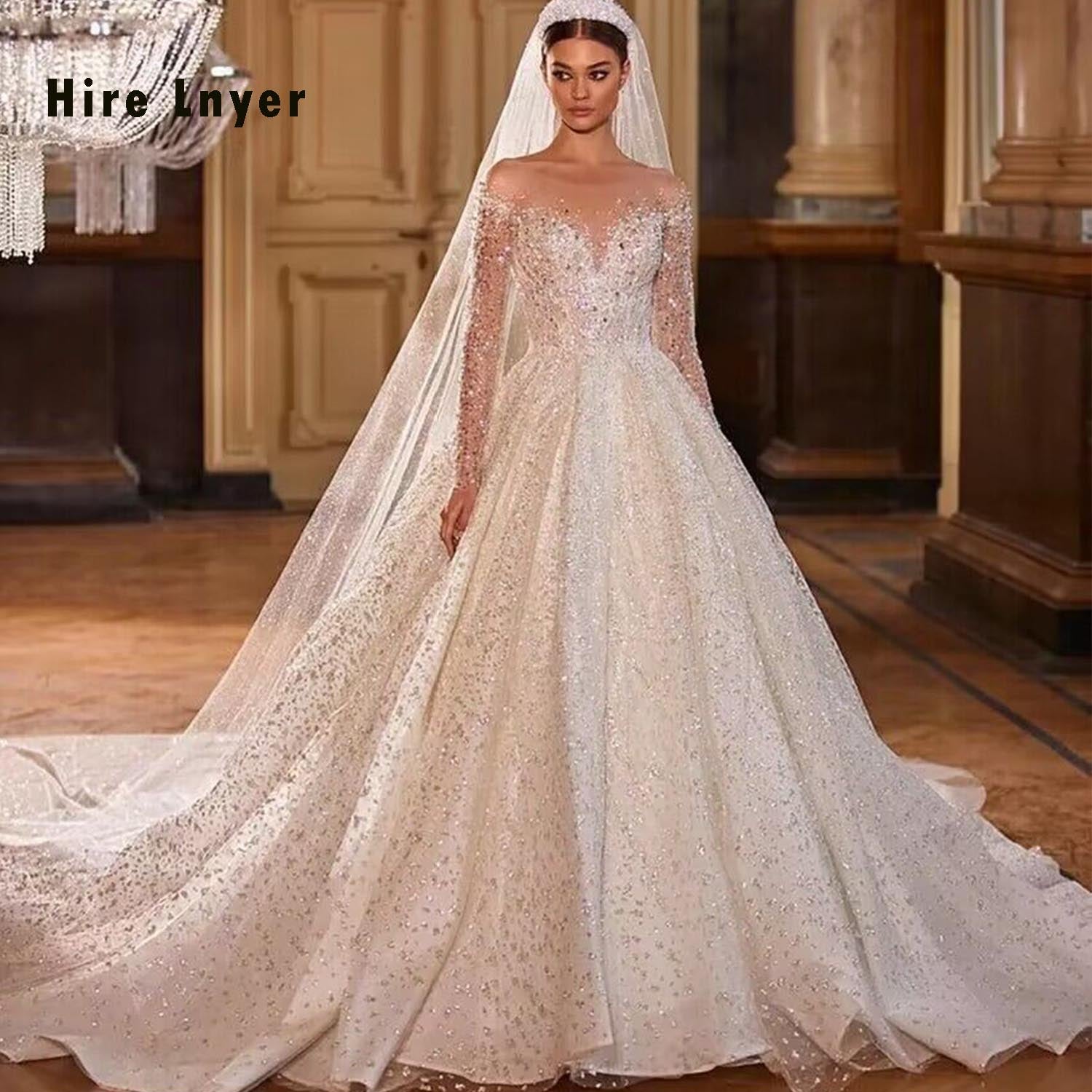 Beautiful bride in an opulent ballroom wearing a crystal beaded shining ball gown wedding dress, featuring sheer long sleeves and a deep plunging illusion neckline.