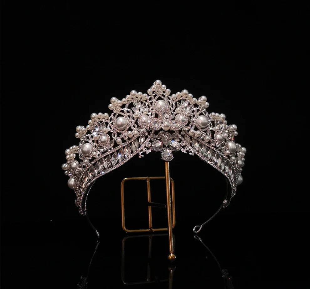 Elegant bridal tiara adorned with pearls and clear crystals, displayed on a gold stand against a black background.