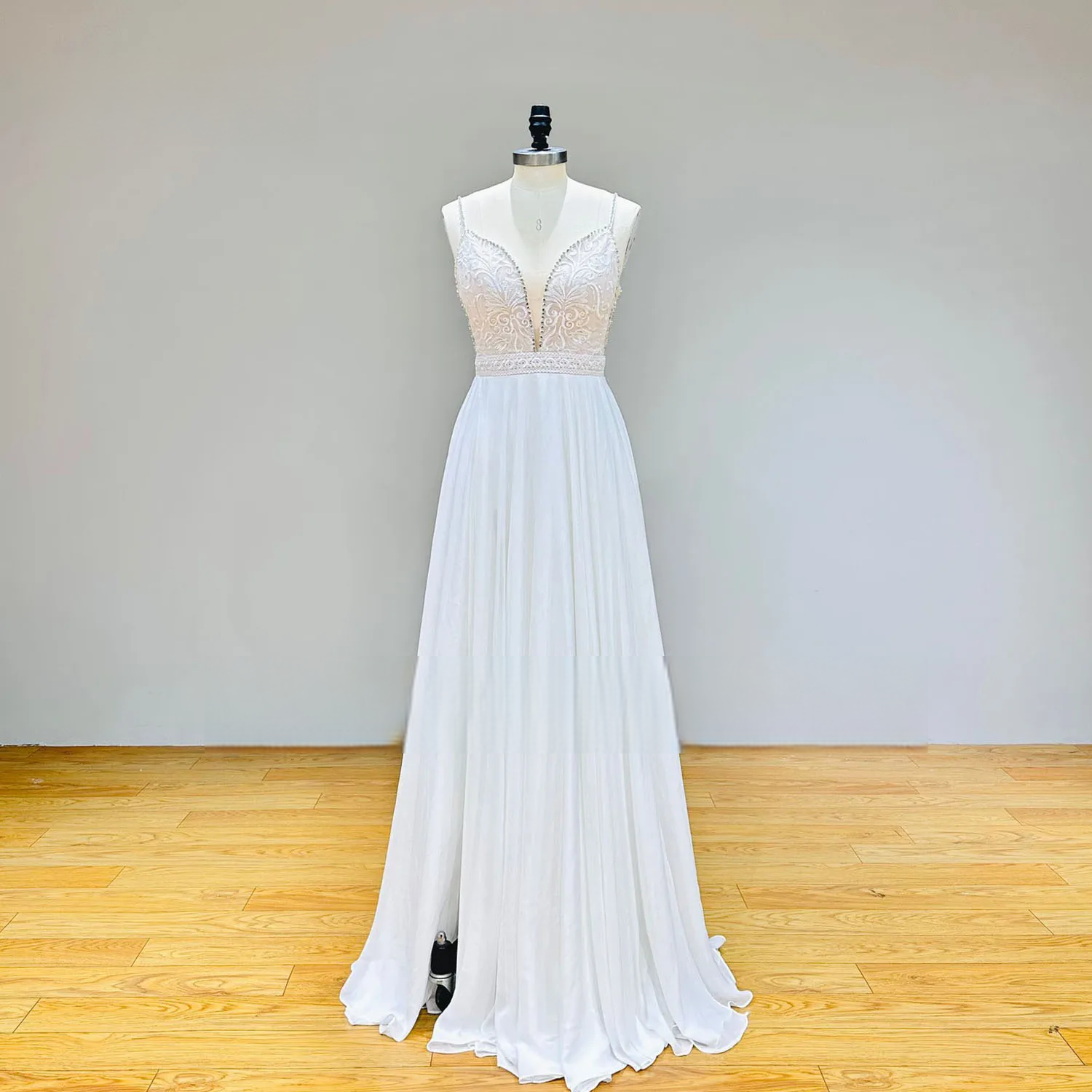 Studio front view of a backless chiffon wedding dress on a mannequin showcasing the plunging V-neckline, beaded lace bodice, and pleated A-line skirt.