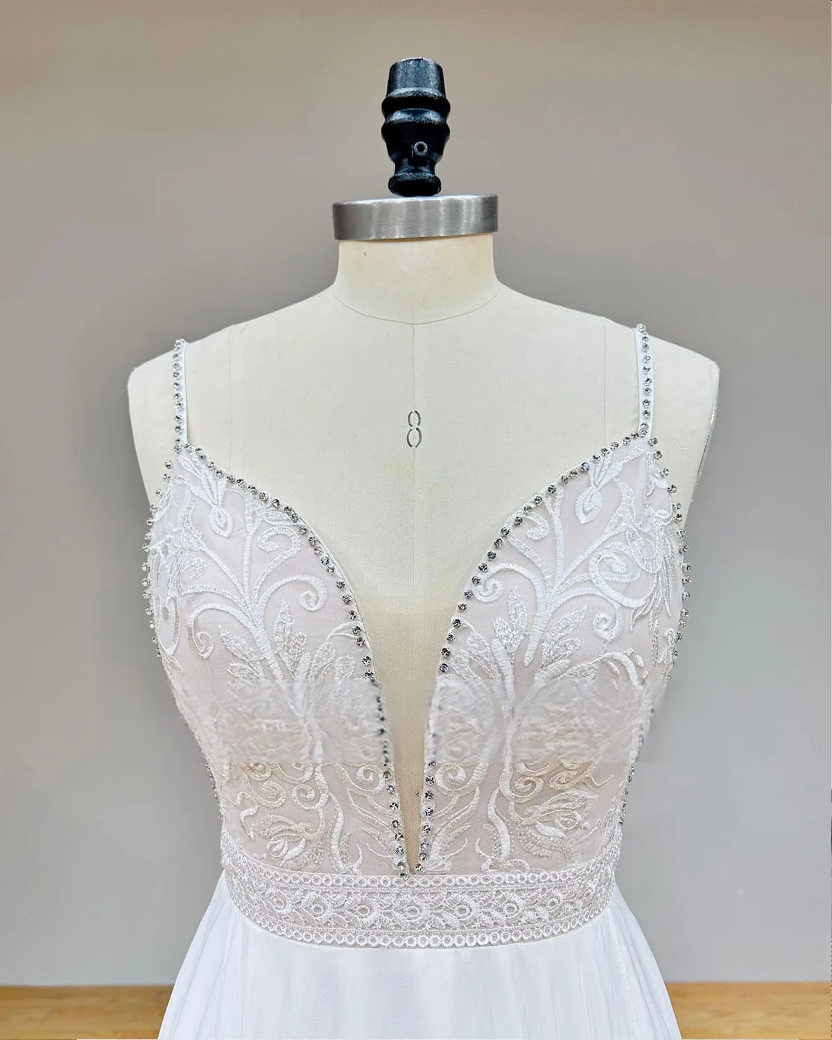 Close-up detail of a bridal bodice featuring intricate white floral lace, a deep plunging V-neckline, and shimmering crystal-embellished spaghetti straps.