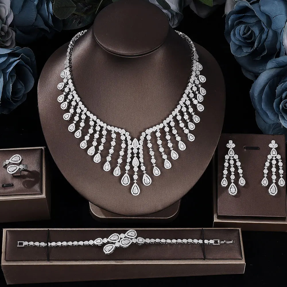 Luxury 4-piece cubic zirconia wedding necklace set presented on a dark brown display bust with matching drop earrings, a link bracelet, and a statement ring alongside blue roses.