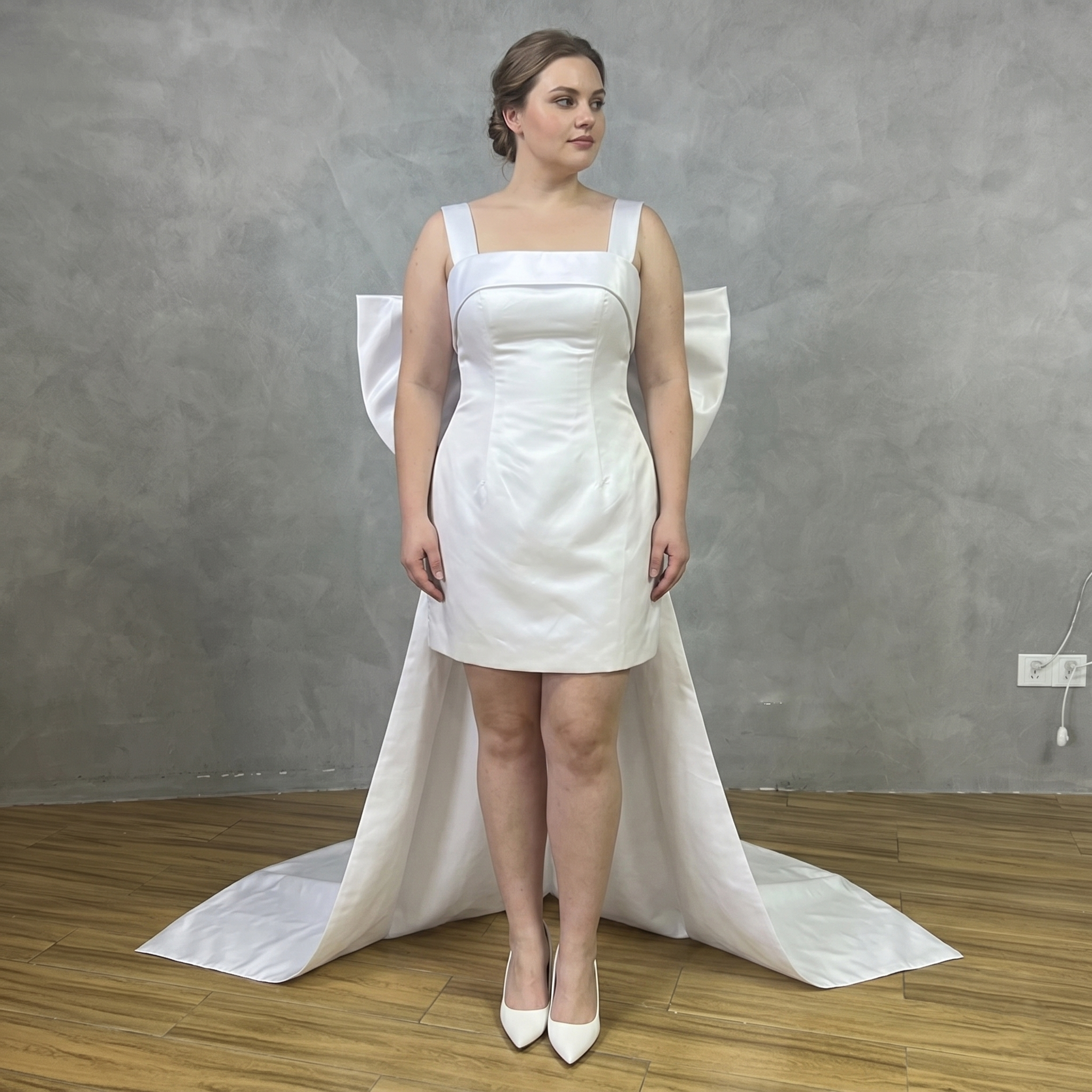 Real bride modeling a sleeveless short white satin bridal gown with a structured square neck bodice and a large statement back bow.