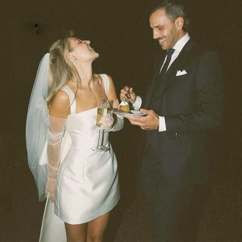 Bride wearing a sleeveless square neck short satin wedding dress with sheer bridal gloves and a tulle veil, laughing during a reception cake cutting ceremony.