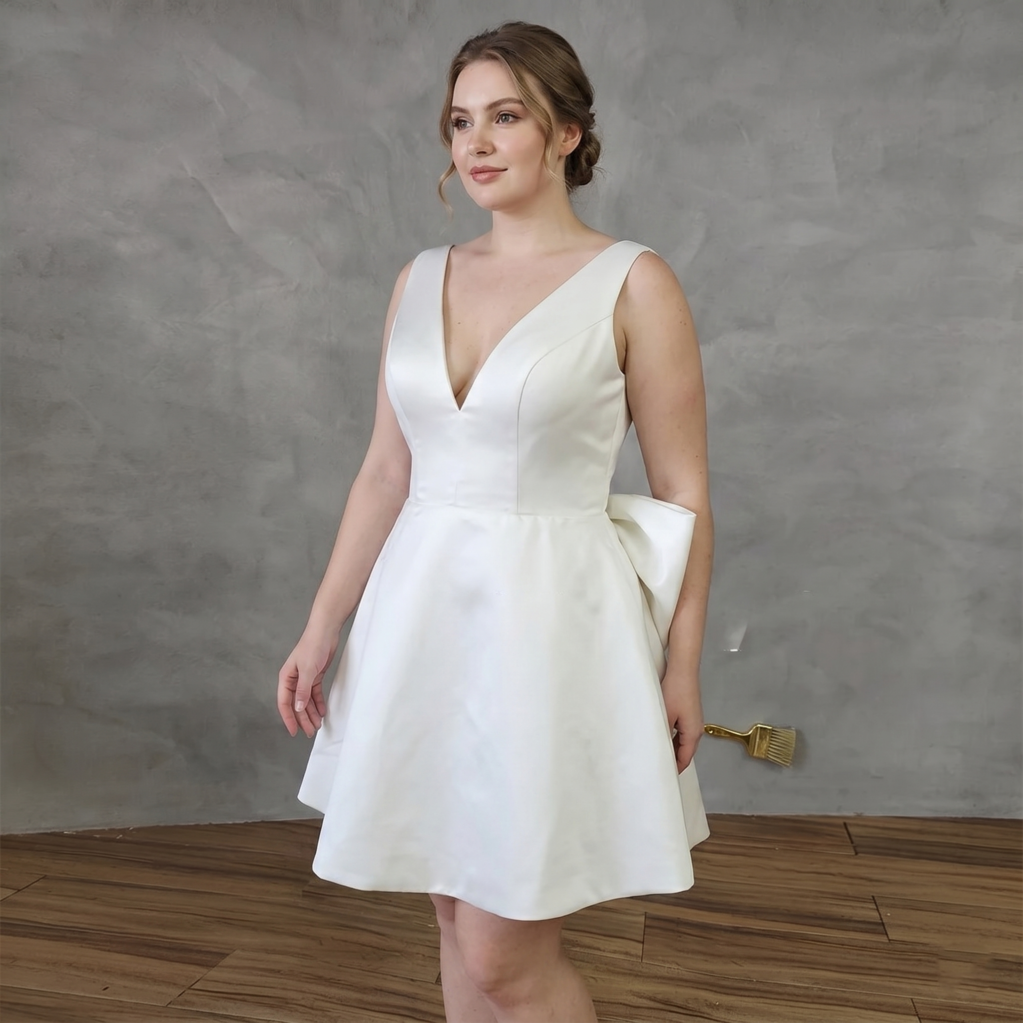 Real bride modeling a sleeveless short white satin bridal gown with a deep V-neckline, structured princess seams, and a large architectural statement bow visible at the back.