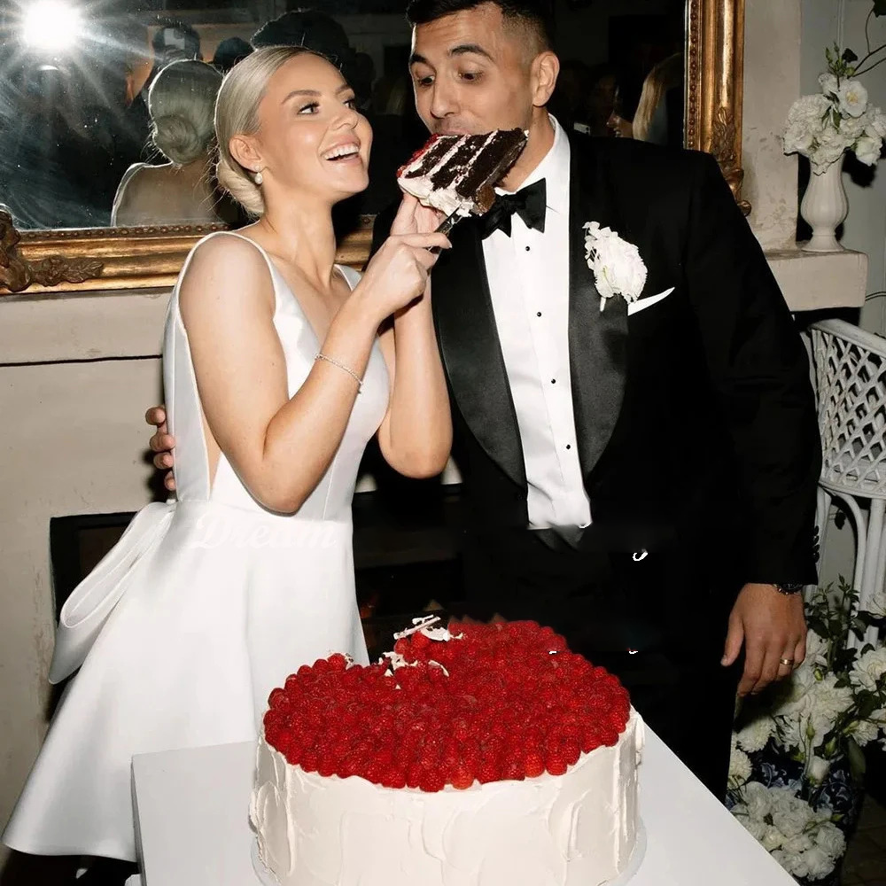 Bride wearing a sleeveless backless short satin wedding dress with a big back bow during a reception cake cutting ceremony, styled with a classic low bun hair look.