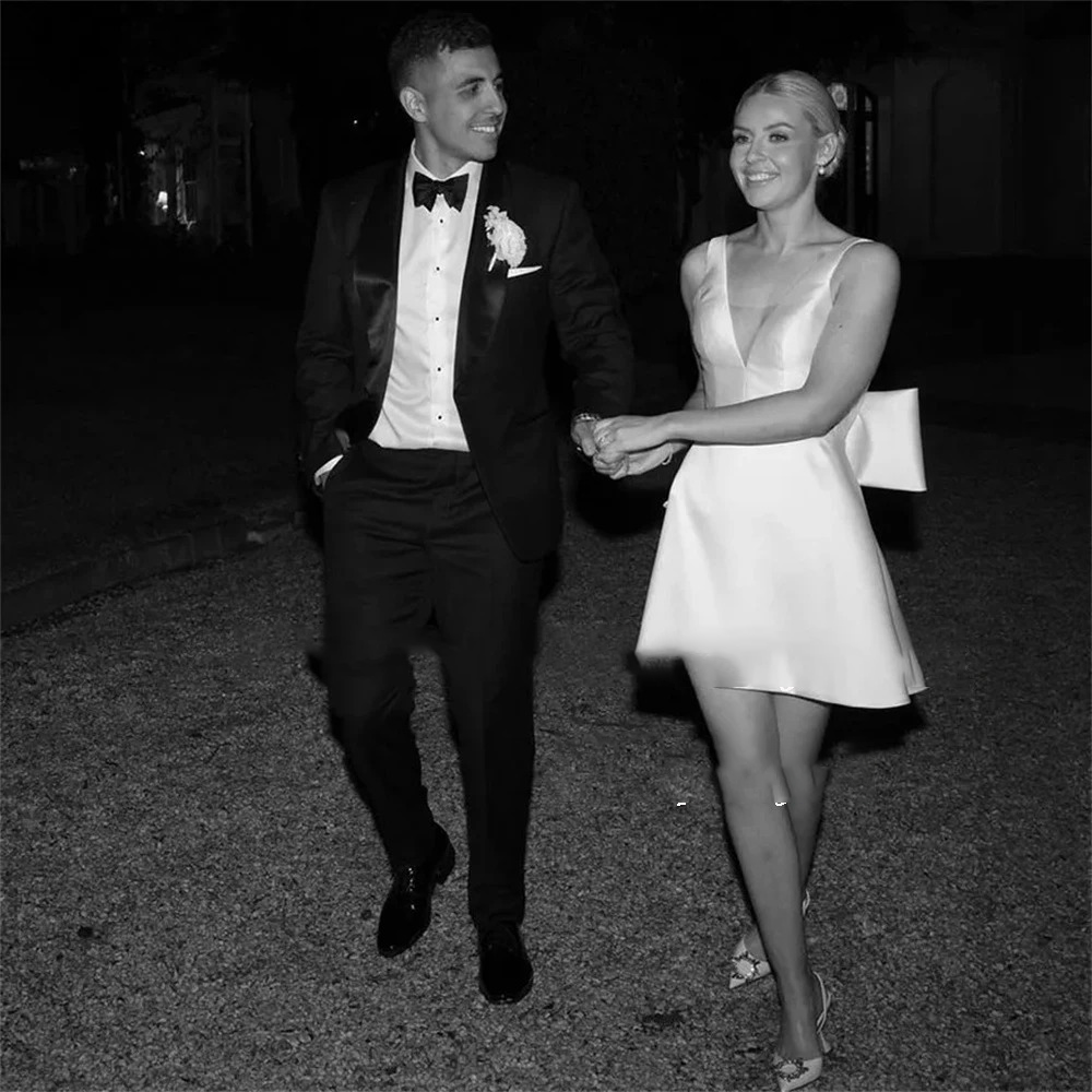 Black and white snapshot of a bride in a short satin reception dress with a plunging V-neckline and detachable back bow, walking hand-in-hand with the groom.