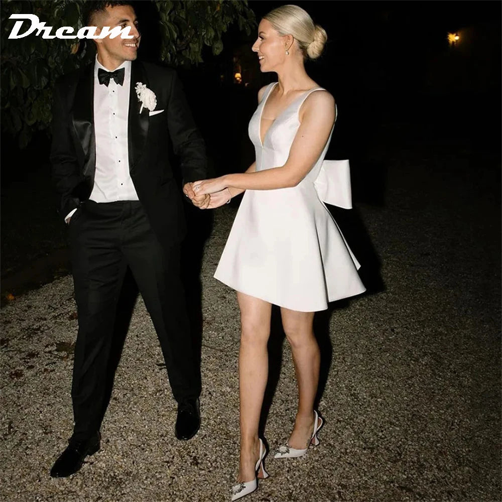 Outdoor evening photo of a short white satin A-line bridal mini dress with a plunging sweetheart illusion top and a structured bow, paired with embellished stiletto pumps.