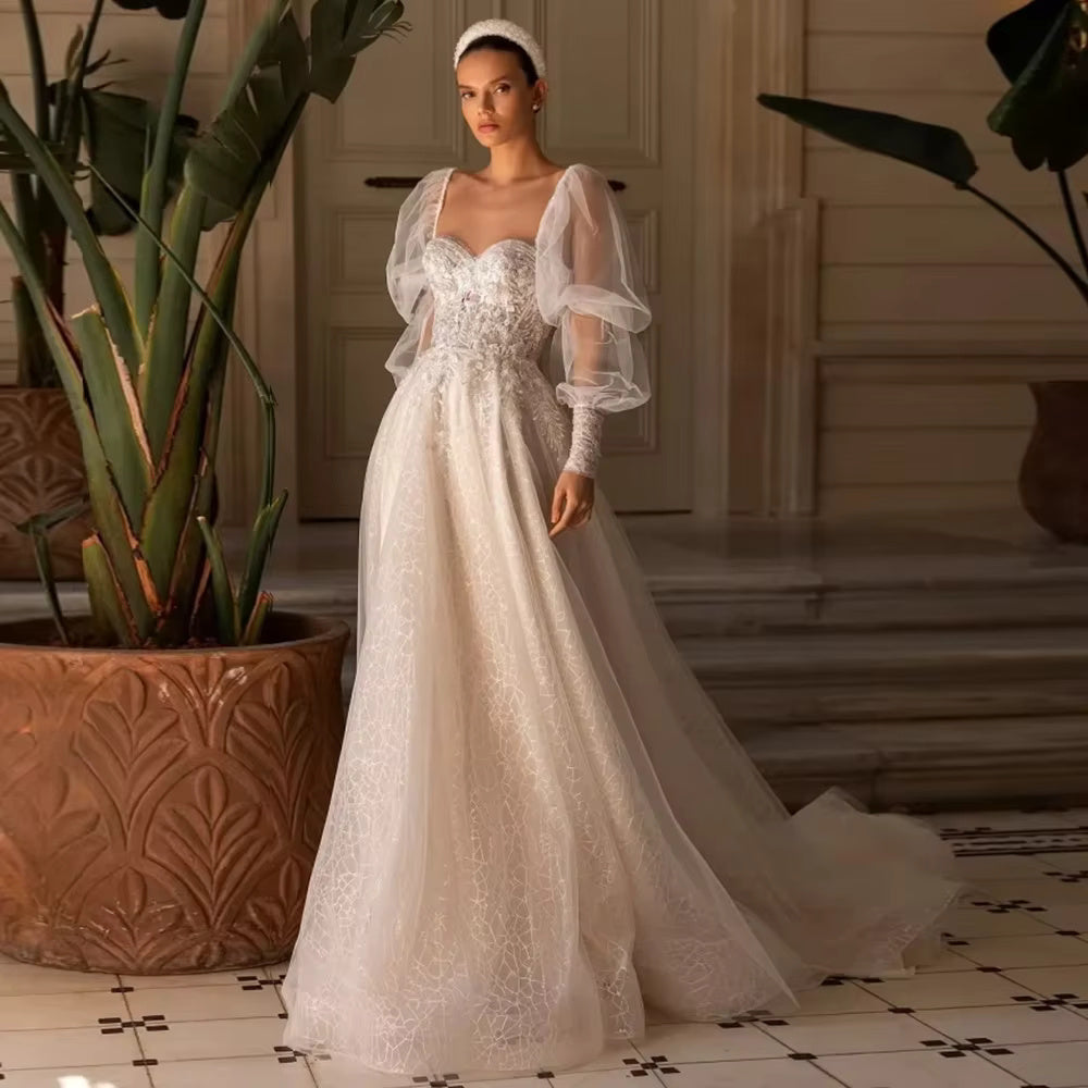 Full-length front view of a bride wearing a romantic tulle wedding dress with a sweetheart neckline, a heavily beaded lace applique bodice, and voluminous detachable sheer long puff sleeves.