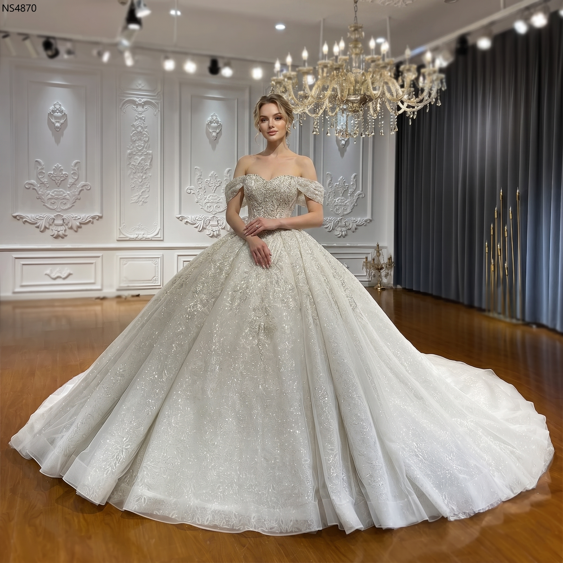 Full-length front view of an elegant model wearing a luxury ball gown wedding dress with detachable off-the-shoulder straps and a shimmering, heavily beaded bodice in a grand showroom setting.