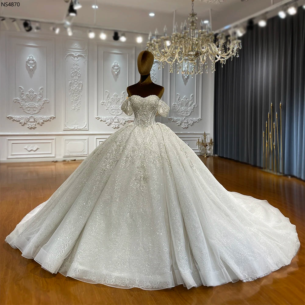 Showroom display of a strapless luxury wedding dress on a mannequin, highlighted by elegant detachable ruffled sleeves and a majestic, sparkling cathedral-length skirt.