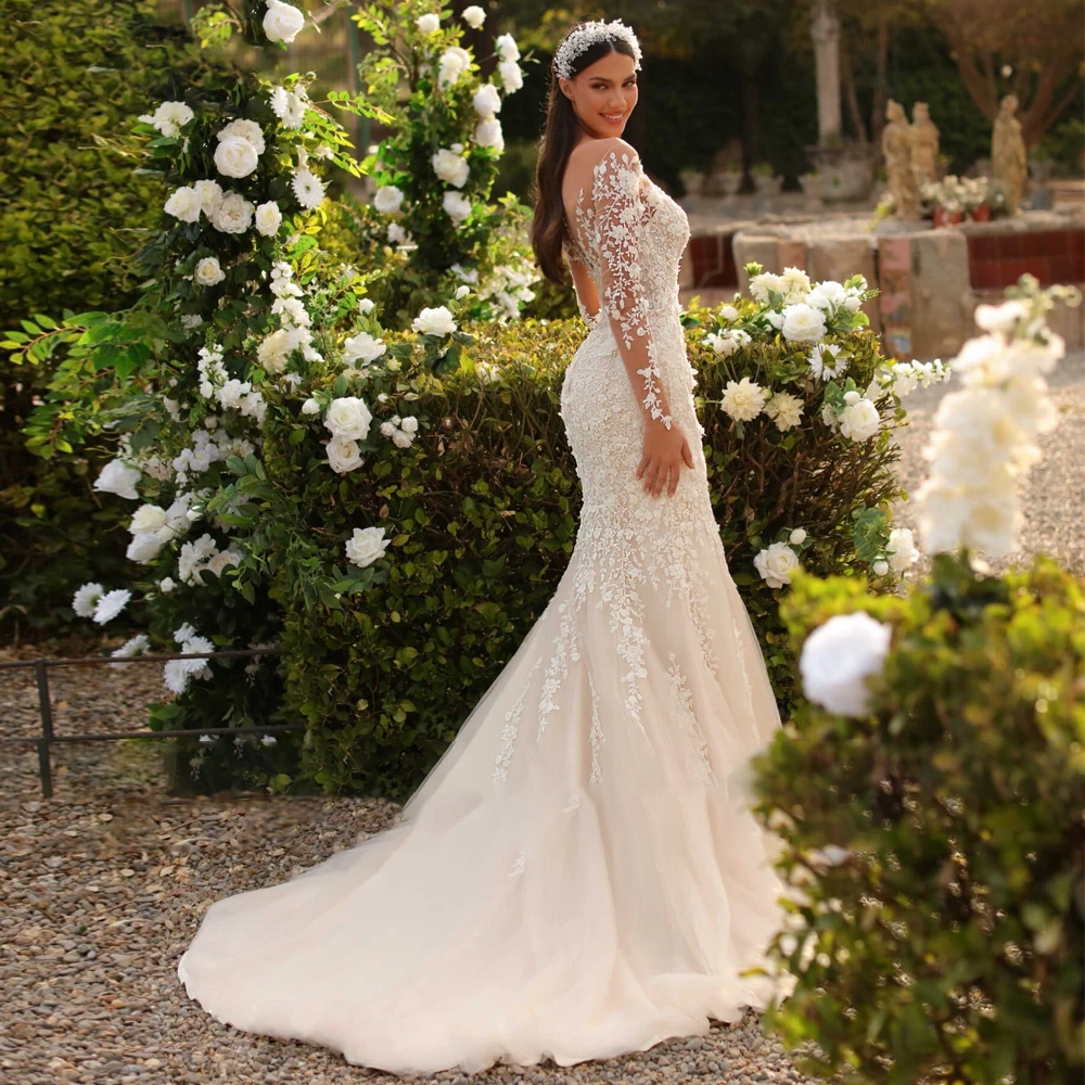 A side view of a bride posing in a mermaid-style bridal gown, highlighting the lace embroidery, sheer sleeves, and detachable train in a garden setting.