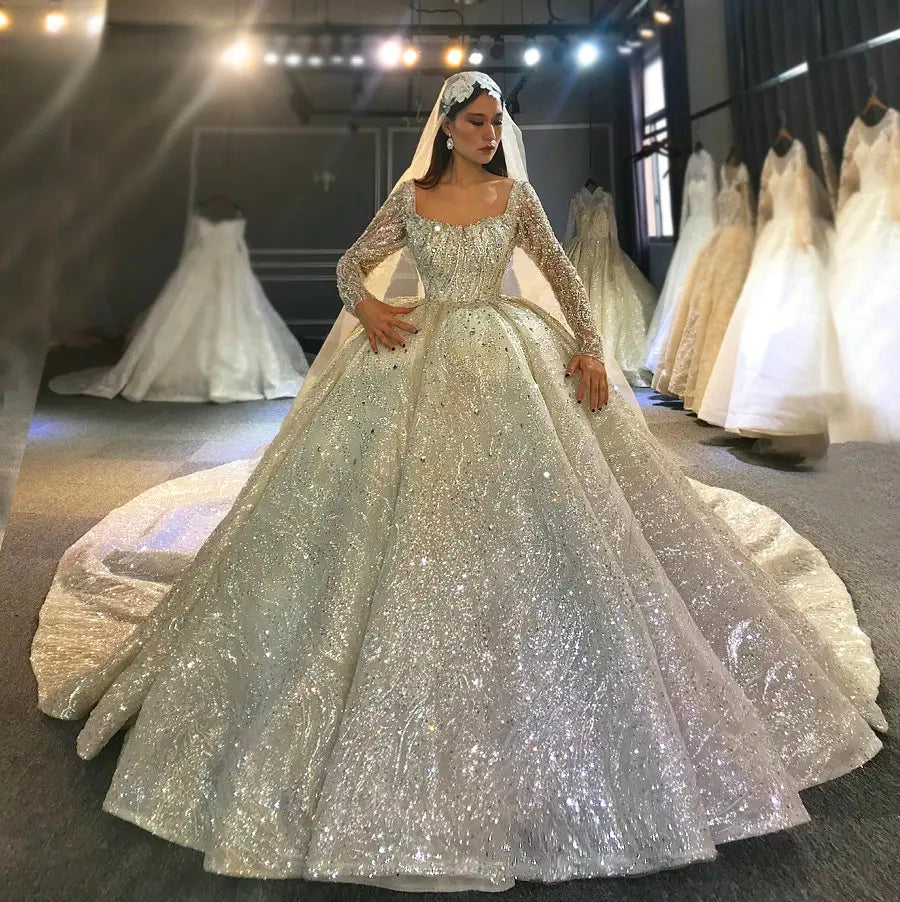 Full view of a shimmering Dubai princess luxury beaded wedding dress featuring long sleeves, a square neckline, and a voluminous ball gown skirt in a bridal showroom.