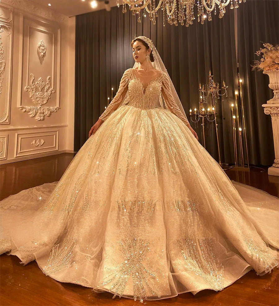 Full-length front view of a Dubai style royal train ball gown wedding dress featuring shimmering gold-tone beadwork and long sheer sleeves in a luxury bridal showroom.