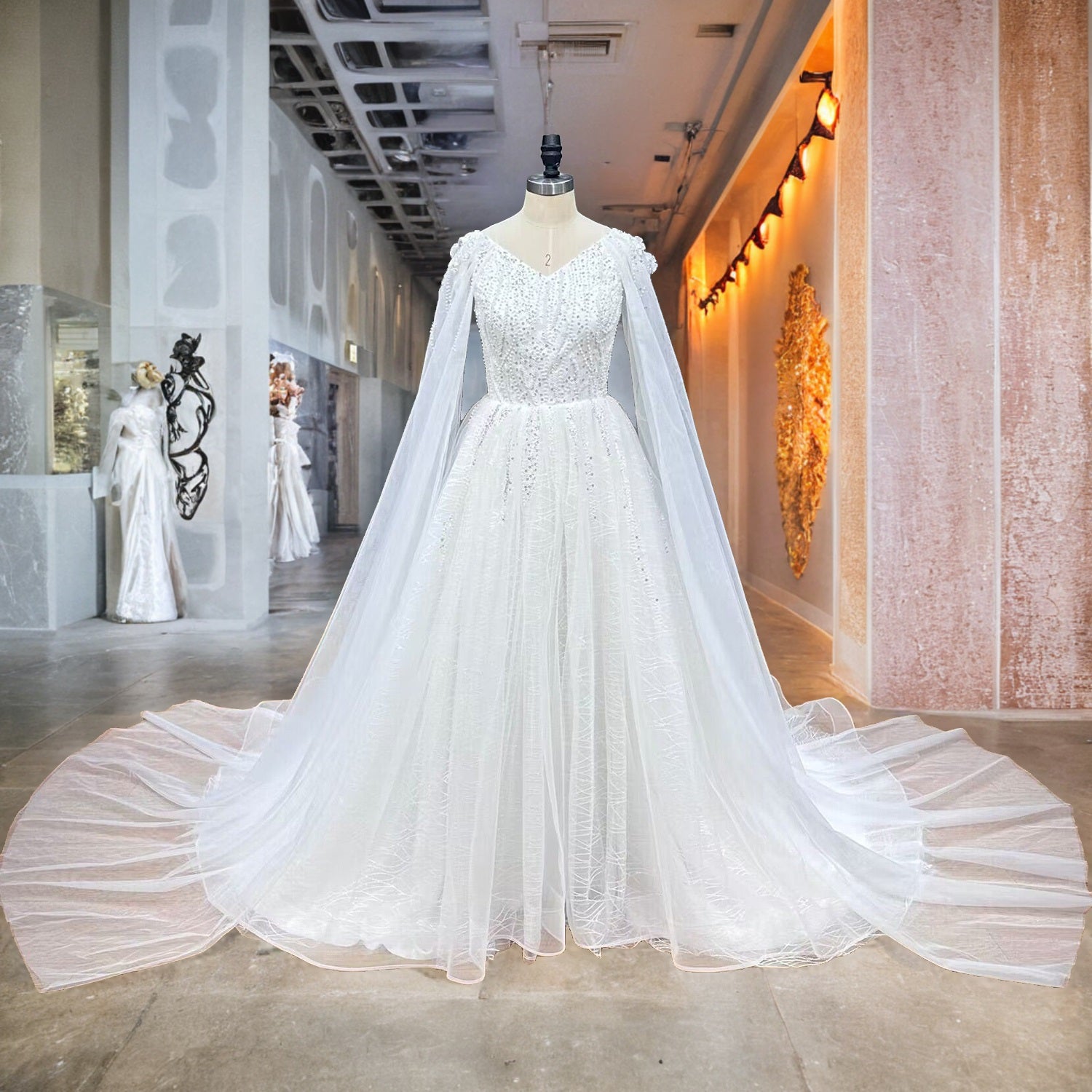 Front full-length view of a modern A-line wedding dress with a pearl-beaded bodice and cape sleeves on a showroom mannequin.