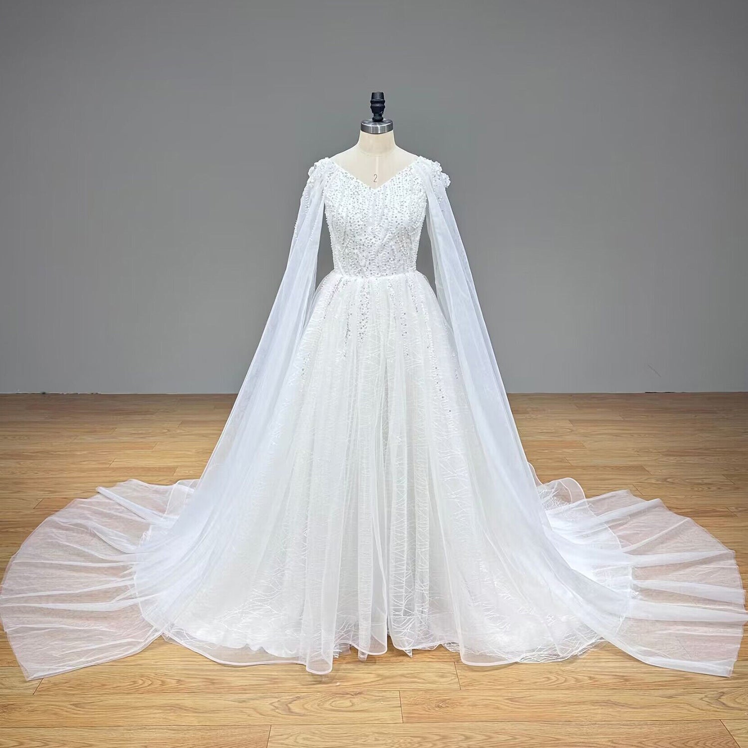 Studio product shot of a luxury V-neck bridal gown featuring a shimmering tulle skirt and elegant shoulder cape drapes.