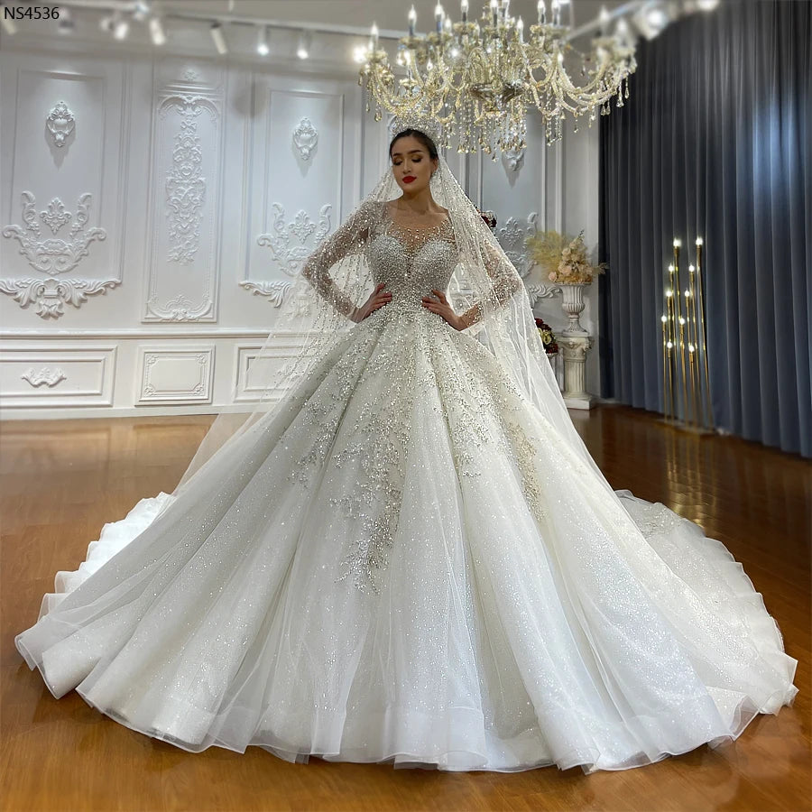 Full-length front view of an elegant model in a luxury long-sleeve ball gown wedding dress featuring intricate pearl beading and a voluminous sparkling tulle skirt.
