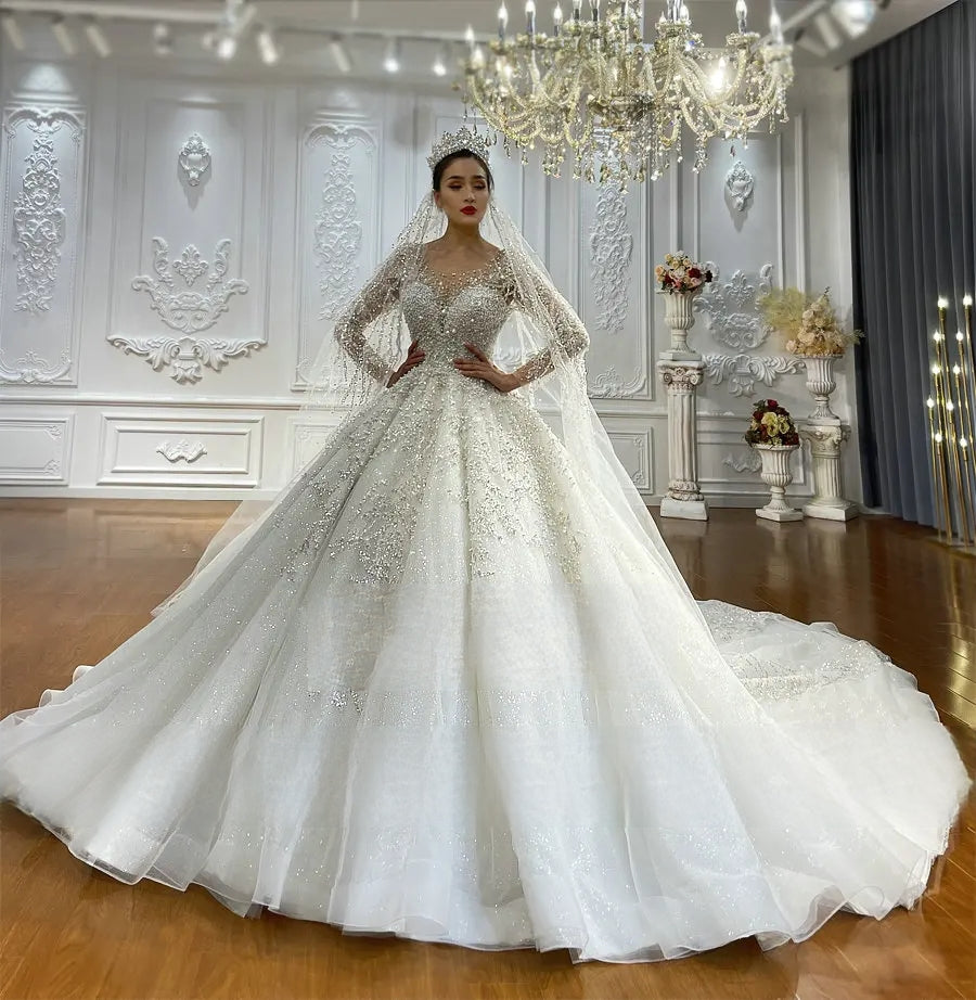 Elegant front perspective of a shimmering A-line bridal gown with long sleeves and a regal ballroom silhouette, displayed under a crystal chandelier in a bridal boutique.