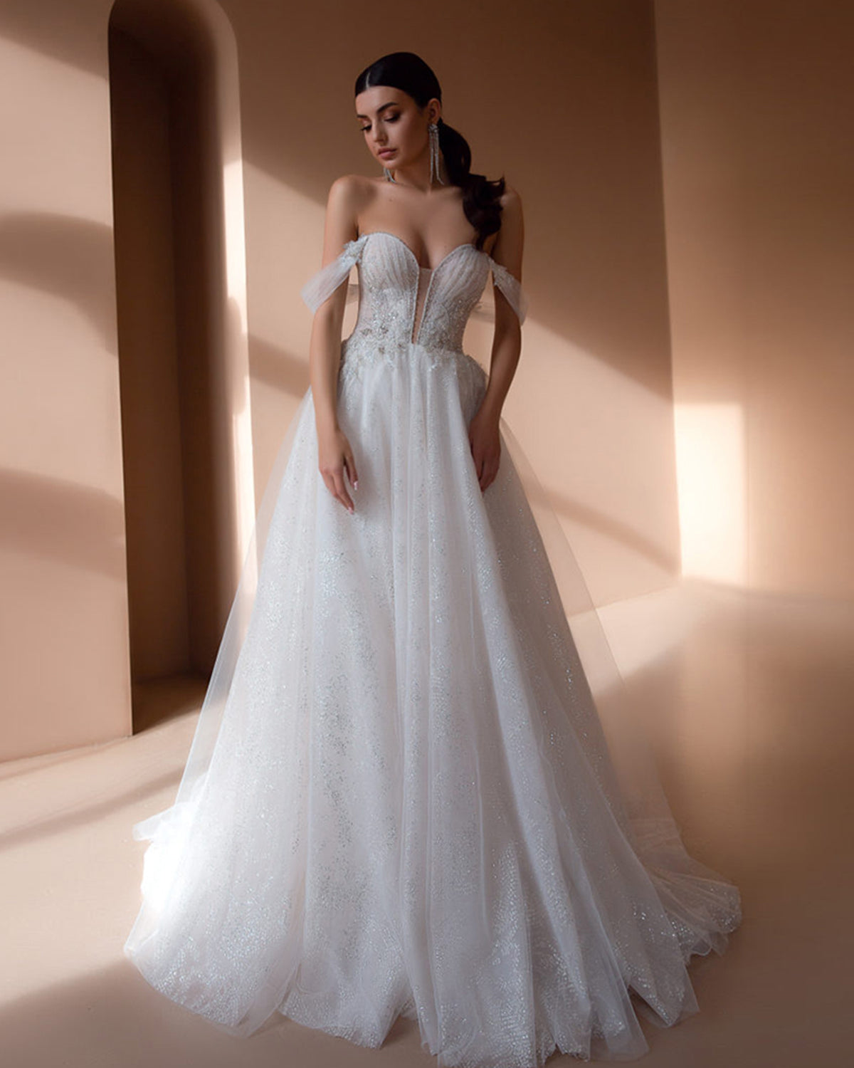 Smiling bride wearing an elegant off-shoulder A-line wedding dress with a heavily beaded bodice, holding a pastel bouquet under a romantic rose-covered floral arch.