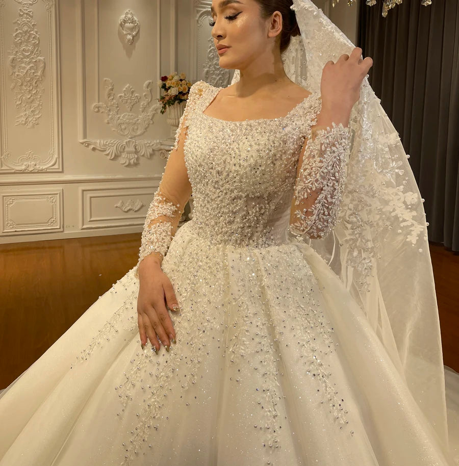 Close-up detail of a bridal gown bodice featuring a square neck, illusion long sleeves, and heavy pearl and bead embellishments for a luxurious texture.