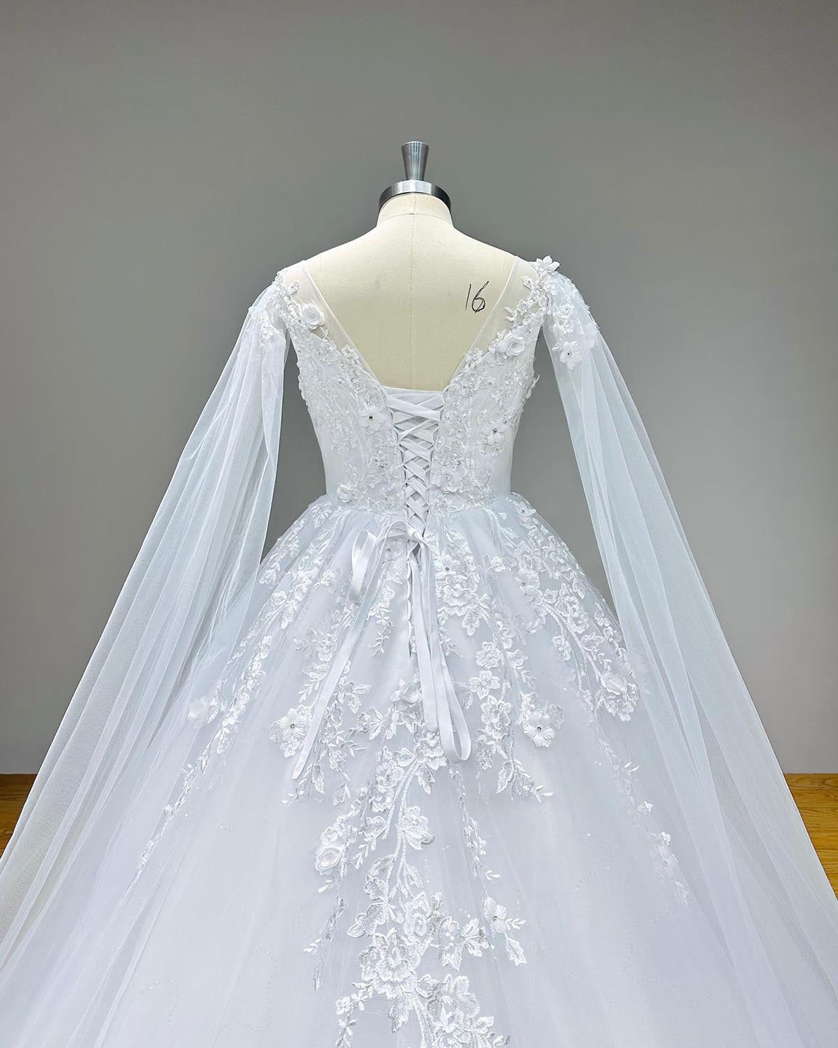 Close-up back bodice detail of an elegant backless wedding dress on a mannequin, showcasing the sheer V-illusion back, traditional lace-up corset tie, and delicate floral appliques where the long shawl sleeves attach.