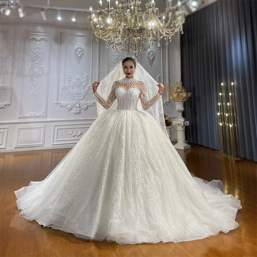 Full-length front view of an elegant beaded ball gown wedding dress featuring a high neck, long sleeves, and a shimmering voluminous skirt, showcased in a luxury bridal showroom under a chandelier.