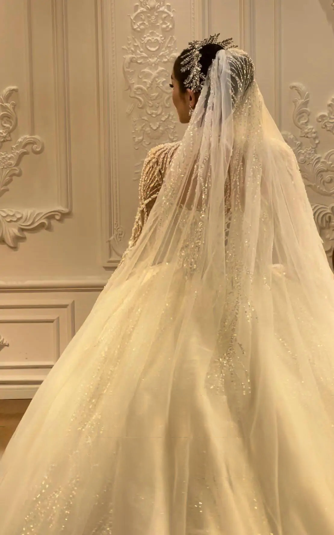Close-up back view of a bride wearing a crystal tiara and a long shimmering veil that complements the intricate beadwork of a long-sleeve luxury wedding gown.