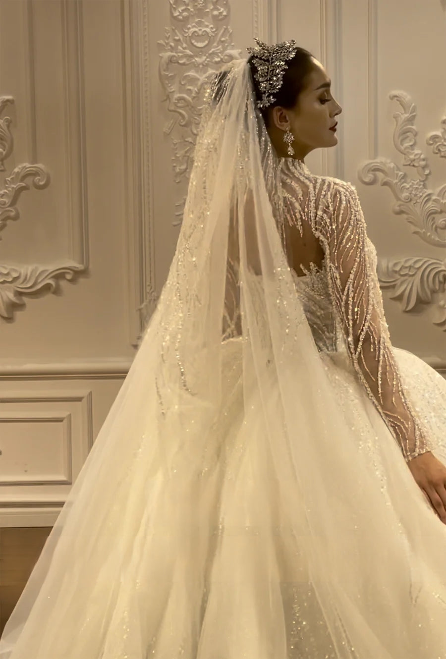 Side profile of a bride in a luxury beaded wedding dress, emphasizing the delicate floral beadwork on the sleeves and the regal silhouette of the crown and veil.
