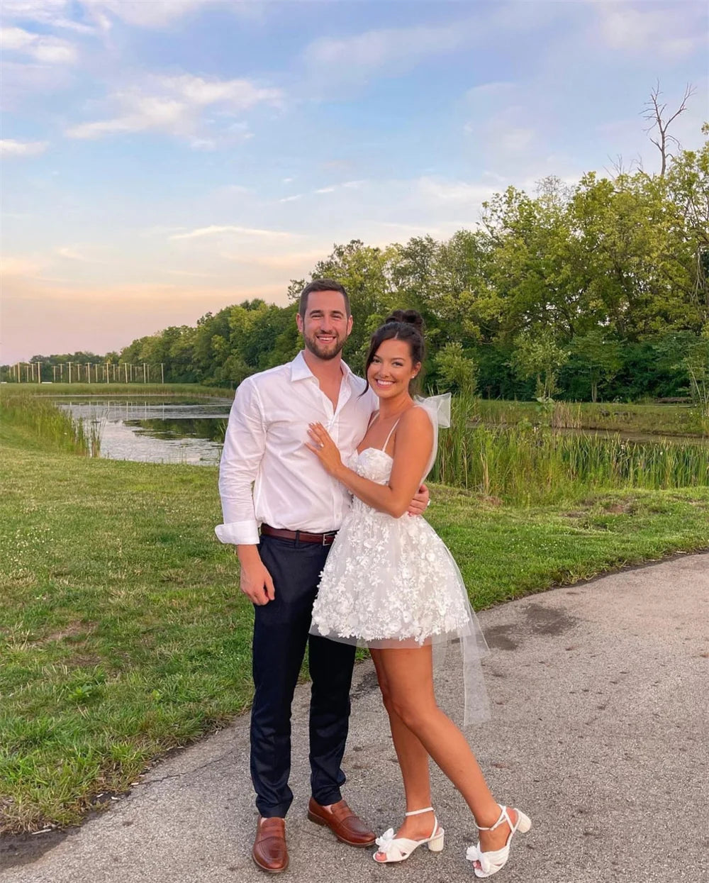 A happy couple in formal attire posing outdoors, with the bride wearing a white floral lace mini dress with a sweetheart neckline.