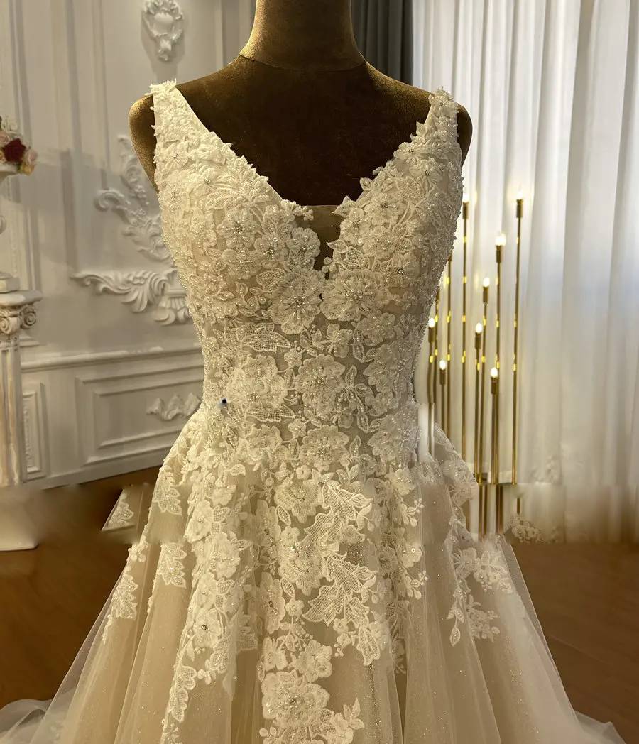 Close-up detail of the bodice of a champagne wedding dress, highlighting the V-neckline and intricate white floral lace applique.