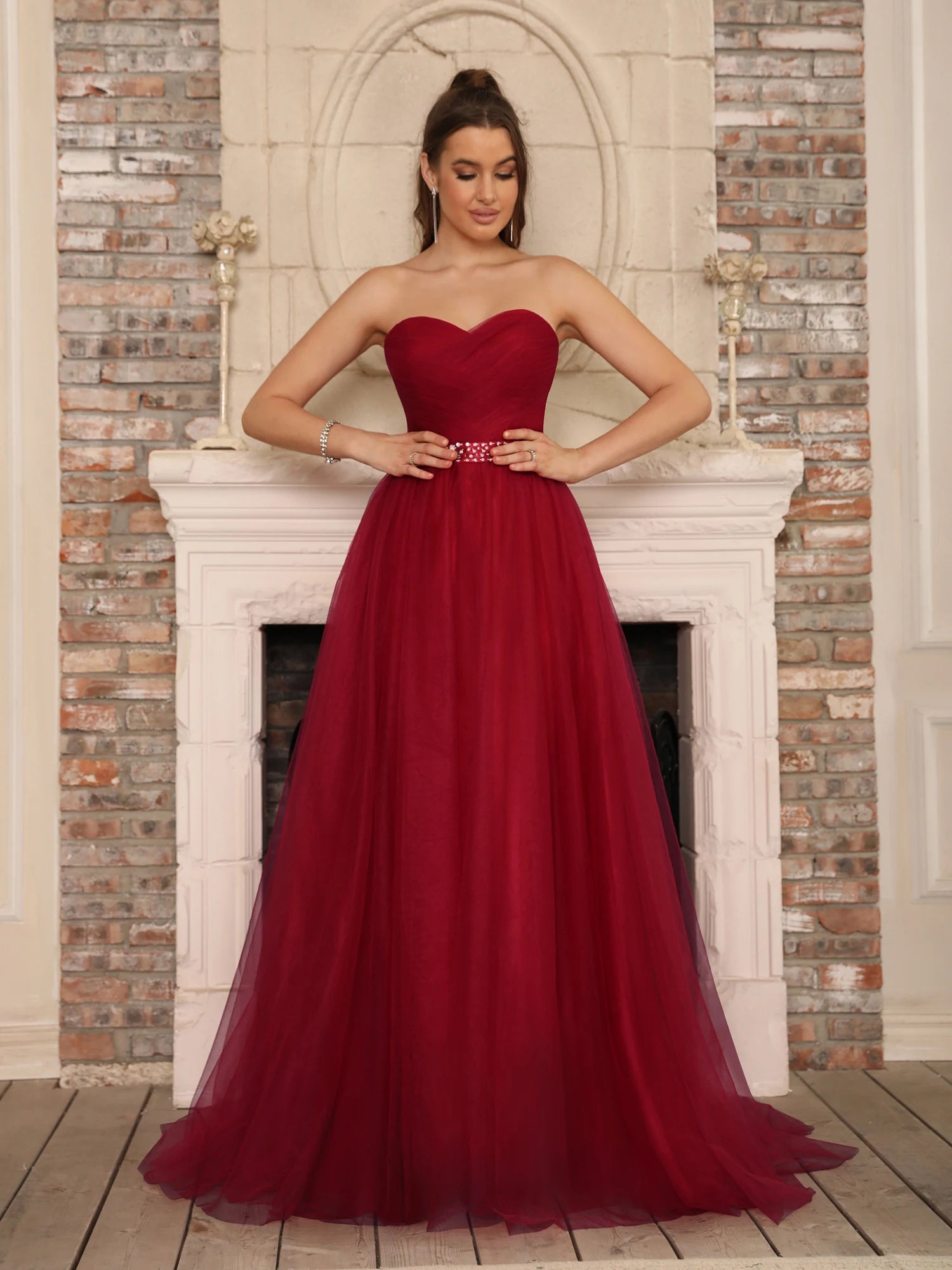 Front view of a burgundy sweetheart neck A-line gown with a pleated bodice and a decorative rhinestone waist belt standing by a fireplace.