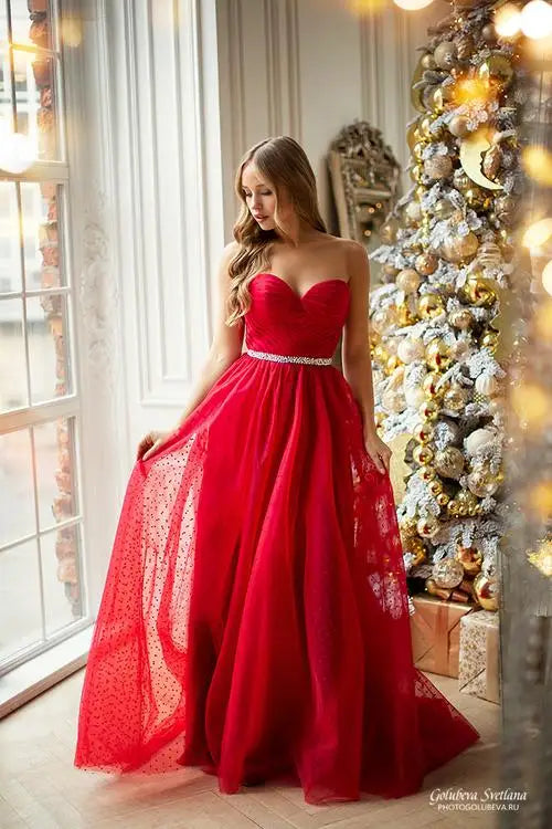 Festive red A-line sweetheart neck gown with a crystal belt, posing in a holiday setting next to a white Christmas tree.