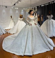 Full view of an elegant full beaded lace wedding dress with graceful trumpet sleeves, a sweetheart neckline, and an expansive sparkling ball gown skirt in a bridal showroom.