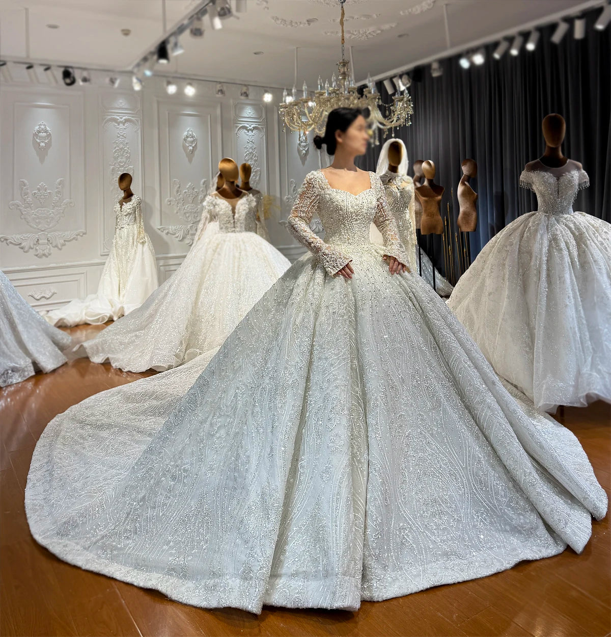 Full view of an elegant full beaded lace wedding dress with graceful trumpet sleeves, a sweetheart neckline, and an expansive sparkling ball gown skirt in a bridal showroom.