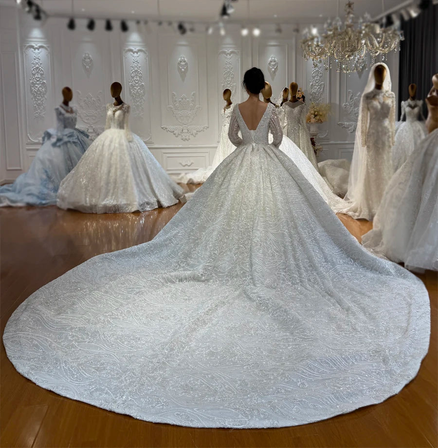 Rear view of a model in a luxury fully beaded wedding dress, displaying the deep V-back design and a massive sparkling lace train spread across the showroom floor.
