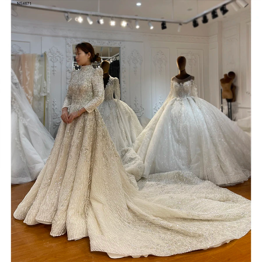 Front perspective of an elegant Muslim bridal gown in a boutique, featuring modest long sleeves, a high neck, full crystal beading, and a sweeping, textured ball gown train.