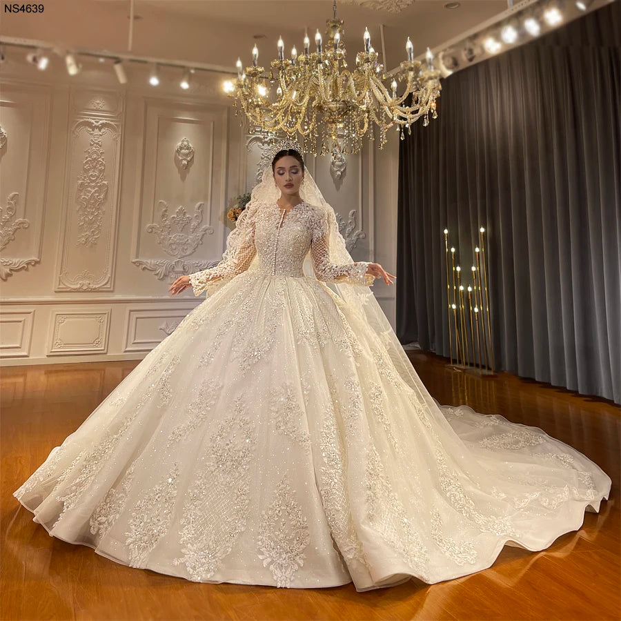 Full view of an elegant full lace ball gown wedding dress with a modest high neckline, long sleeves, and an expansive shimmering skirt, showcased in a luxury bridal showroom.