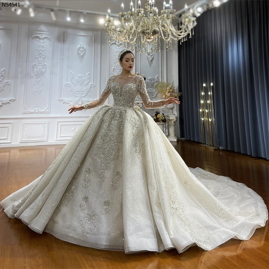 Full-length front view of a model wearing an elegant vintage ball gown wedding dress with a high neckline, long lace sleeves, and a voluminous sparkling skirt.