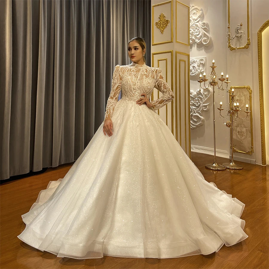 Side profile of a model showcasing a luxury long-sleeved A-line bridal gown with a shimmering sparkly skirt and a detailed beaded top.