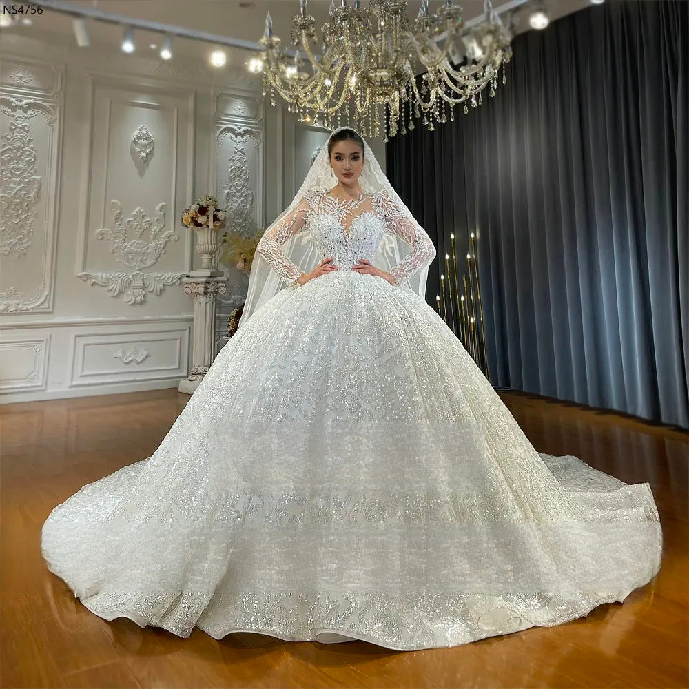 Full-length front view of a luxury shimmering ball gown bridal dress with intricate lace embroidery and long sleeves, showcased under a grand chandelier in a bridal showroom.