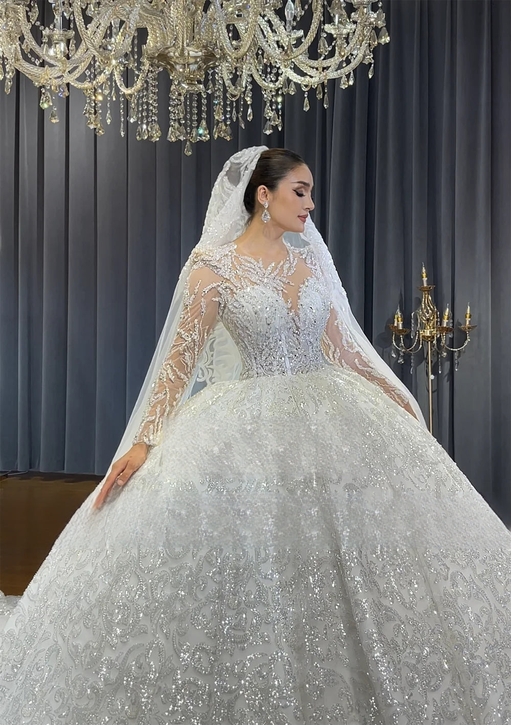 Side profile of a model wearing a luxury sequined ball gown wedding dress with a long train and an elegant lace-edged bridal veil in a boutique setting.