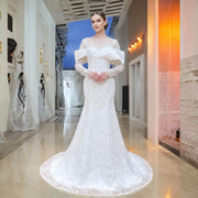 Elegant model posing in a luxury showroom wearing a white lace mermaid wedding dress with long sleeves, a delicate O-neck illusion, and a modern satin bodice drape.