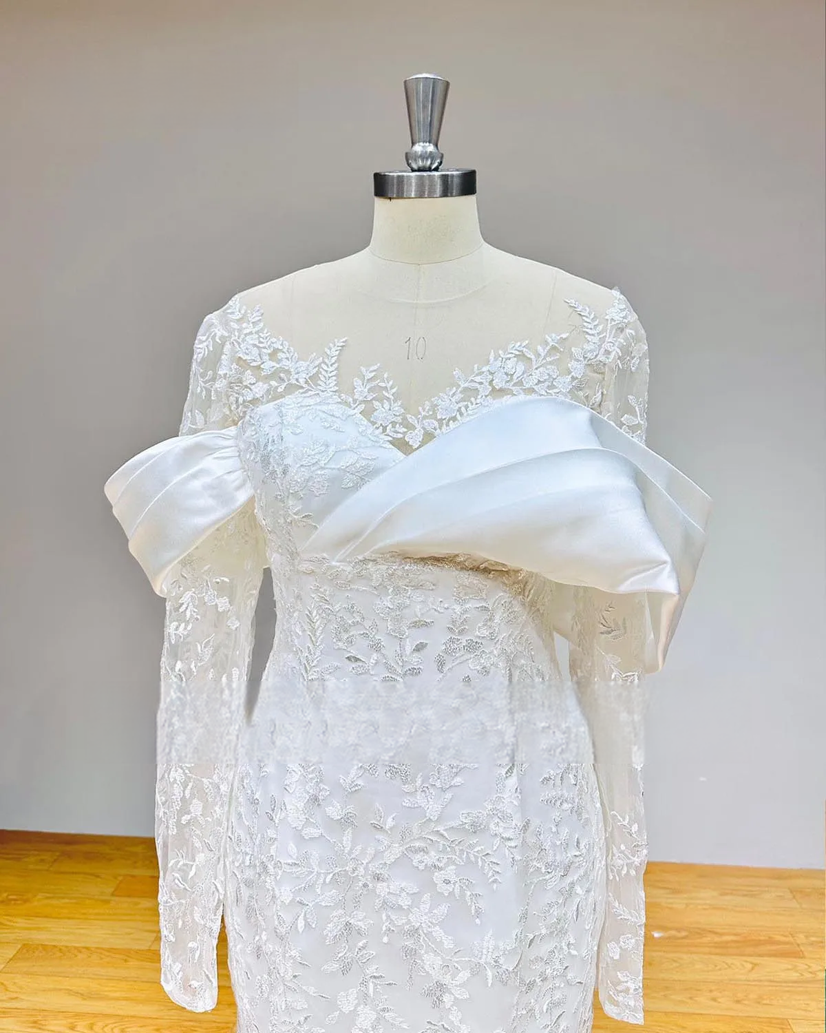 Detailed close-up of the bodice on an elegant lace mermaid wedding dress, showcasing the sheer O-neck, long lace sleeves, and dramatic draped satin off-the-shoulder detail.