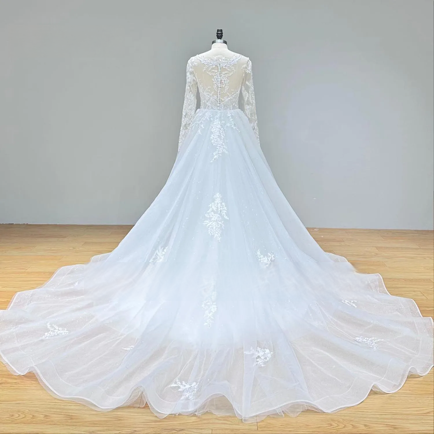 Back view of an elegant long sleeve mermaid wedding dress on a mannequin, detailing the sheer illusion back, row of buttons, and the sweeping detachable train.