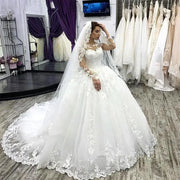 Full-length front view of a model wearing an elegant plus size wedding dress featuring a sweetheart neckline, intricate floral lace appliques, sheer long sleeves, and a flowing layered tulle A-line skirt.