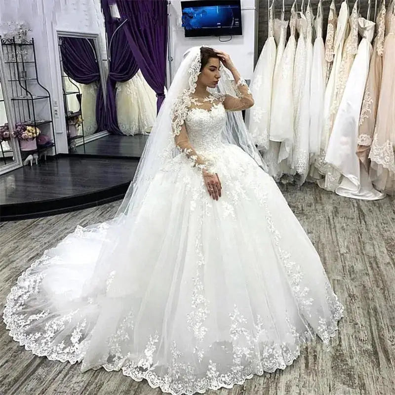 Full-length front view of a model wearing an elegant plus size wedding dress featuring a sweetheart neckline, intricate floral lace appliques, sheer long sleeves, and a flowing layered tulle A-line skirt.