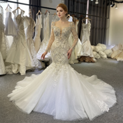 Front view of a bride wearing an elegant long-sleeved lace mermaid wedding gown featuring heavy crystal beadwork, an illusion plunging neckline, and a voluminous flared train.
