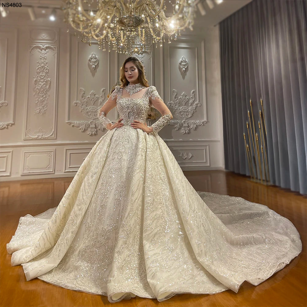 Full front view of a model wearing an elegant luxury ball gown bridal dress featuring a heavily beaded bodice, high-neck illusion collar, long sleeves, and a massive shimmering cathedral train.