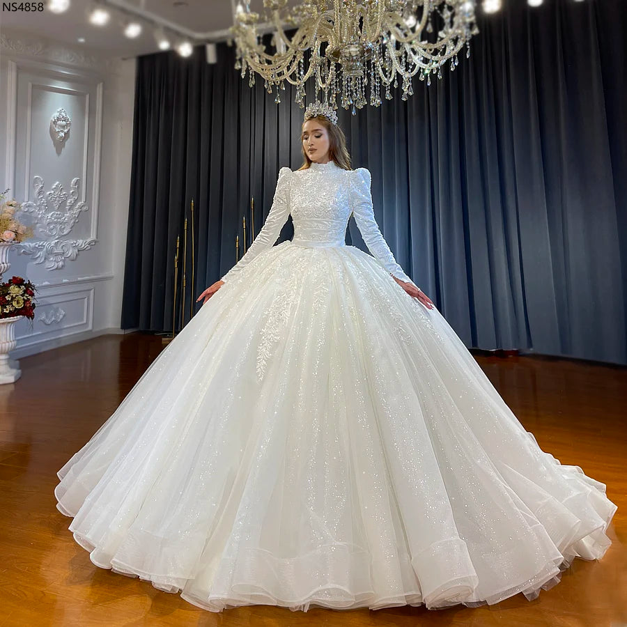 Full front view of a model in a luxury Muslim bridal dress featuring a high neckline, modest long sleeves, and a shimmering, voluminous ball gown skirt in a bridal boutique.