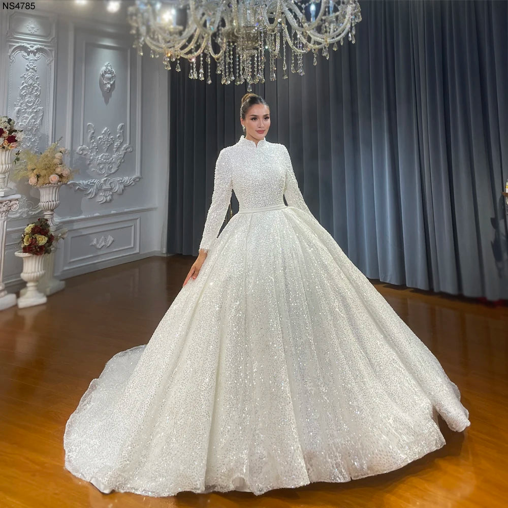 Full front view of a model in an elegant Muslim bridal ball gown with full lining and long sleeves, featuring heavy shimmering sequin embellishments in a luxury showroom.